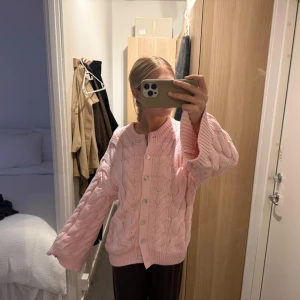 Stickad kofta - Supermysig ljusrosa kofta från Gina Tricot med grovstickat flätmönster och stora vita knappar framtill. Koftan har lång ärm, ribbade muddar och en loose fit som ger en chill vibe. Perfekt att slänga på sig när det är lite kyligt ute. Helt slutsåld!