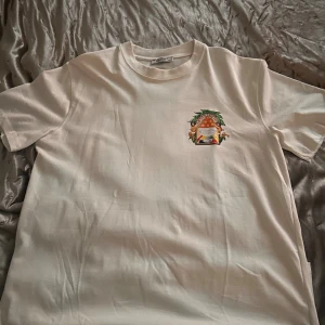 Vit t-shirt från Casablanca med print - Vit t-shirt från Casablanca med färgglatt print på ryggen och mindre tryck på bröstet. Motivet har änglar, apelsiner och texten 'Casablanca'. Klassisk passform och rund halsringning. Tillverkad i mjuk bomull som känns skön mot huden. Storlek xxl men sitter som L