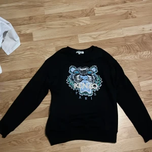 Svart Kenzo tröja med tigertryck - Svart tröja från Kenzo med ikoniskt broderat tigerhuvud och texten 'Kenzo Paris' framtill. Tröjan har rund halsringning och långa ärmar. Perfekt statement piece för dig som gillar streetstyle och vill sticka ut.