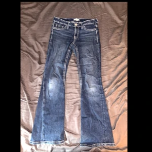Blå bootcut jeans från Gina Tricot - Snygga blå jeans från Gina Tricot i bootcut-modell med ljusa slitningar och kontrastsömmar. Jeansen har klassiska fickor fram och bak med dekorativa broderier på bakfickorna. Perfekt för dig som gillar en retro vibe och vill ha ett par jeans med lite extra attityd.  Midja 39cm  Total längd från midja till ben öppningen 96cm  Bredd på Ben öppningen är cirka 23,6 cm