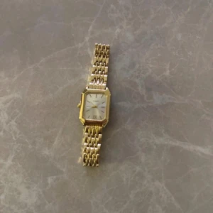 Guldfärgad rektangulär klocka - Snygg guldfärgad klocka från Seiko med rektangulärt ur och länkarmband i metall. Urtavlan är vit med romerska siffror och tunna visare. Perfekt accessoar för dig som gillar klassisk och stilren design.