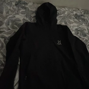 Svart vindjacka från Haglöfs med huva - Svart vindjacka från Haglöfs med minimalistisk design och diskret logga på bröstet. Jackan har huva, långa ärmar och dragkedja framtill. Perfekt för blåsiga dagar och outdoor-äventyr. Snygg och funktionell stil som funkar året runt.Storlek M fast de känns som S