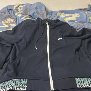 Marinblå hoodie från BOSS med dragkedja - Limited edition Snygg marinblå hoodie från BOSS med vit dragkedja och huva. BOSS-logga på bröstet och upprepade loggor längs nederkanten. Mjukt material och coola gröna detaljer på insidan av kragen. Perfekt för en chill och stilren look.