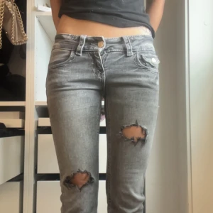 Grå slitna bootcut jeans med broderi - Grå jeans med bootcut passform och slitna hål på båda knäna. Snygga detaljer med broderade bakfickor och silvriga knappar. Jeansen har låg midja och är tillverkade i denim med en tvättad look. 