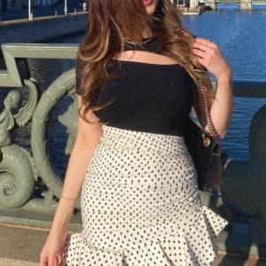 Prickig vit volangkjol - Supersöt vit kjol med svarta prickar och volangkant. Kjolen är kort och har en figurnära passform som framhäver midjan. Perfekt för dig som gillar trendiga detaljer och ett lekfullt uttryck. Passar till både sneakers och klackar.