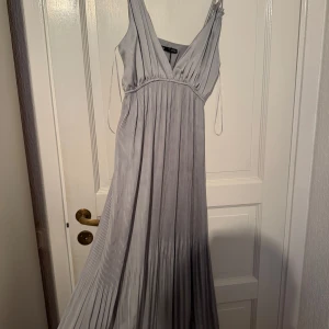Ljusgrå plisserad långklänning - Säljer en elegant ljusgrå långklänning från Zara med djup v-ringning och tunna axelband. Klänningen har plisserad kjol som ger ett snyggt fall och är tillverkad i ett mjukt, glansigt material som känns lyxigt. Perfekt för dig som vill sticka ut med stil.