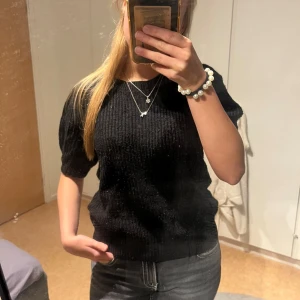 Svart stickad tröja från Vero Moda - Svart stickad tröja från Vero Moda med korta ärmar och rund halsringning. Tröjan har en ribbad struktur och en klassisk, enkel design som är lätt att matcha med olika outfits. Den är lite nopprig… men perfekt för dig som gillar en stilren och bekväm look.😊