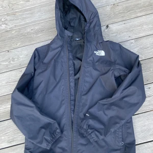 Svart vindjacka från The North Face S - Säljer en svart vindjacka från The North Face i storlek S. Jackan har huva, dragkedja framtill och elastiska muddar vid ärmsluten. Perfekt för blåsiga och regniga dagar och enkel att slänga på sig. Klassisk logga på bröstet och ryggen.