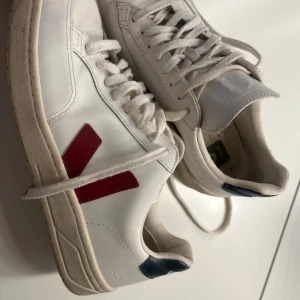 Vita Veja sneakers med röd detalj - Säljer ett par klassiska Veja sneakers i vitt skinn med röd V-logga på sidan och mörkblå detalj vid hälen. Skorna har snörning och platt sula, perfekt för en clean och stilren look. Storlek 40.