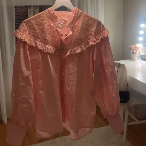 Rosa volangskjorta  - Supersöt ljusrosa skjorta från H&M i bomull med broderade detaljer och volang längs axlarna i storlek XS. Skjortan har långa ärmar, knappar framtill och ett unikt hålmönster på axlar och ärmar. Perfekt för dig som gillar romantiska och trendiga plagg. Hör gärna av er vid frågor eller funderingar! 💖