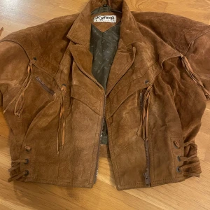 Brun mockajacka med fransar från Jofama - Säljer en brun mockajacka från Jofama med snygga fransdetaljer och snörning i sidorna. Jackan har dragkedja framtill, klassisk krage och långa ärmar med dragkedjor vid ärmsluten. 