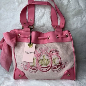 Juicy Couture daydreamer tote rosa axelväska med rosett - Superfin axelväska från Juicy Couture i rosa och ljusrosa mocka och syntet. Väskan har broderad krona och JC-logga framtill, stora handtag, snygga gulddetaljer och en stor mjuk rosett i sammet på sidan. Dragskostängning upptill och matchande nyckelring medföljer.