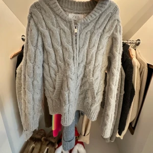 Ljusgrå stickad kofta från Zara - Supermysig ljusgrå stickad kofta från Zara med grovt kabelstickat mönster. Koftan har dragkedja framtill, två fickor och ribbade muddar. Perfekt för lager-på-lager och chill dagar. Nopprig!! 