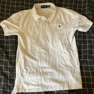 Vit pikétröja från Polo Ralph Lauren - Vit pikétröja från Polo Ralph Lauren med klassisk krage och korta ärmar. Tröjan har broderad logga på bröstet och är i bomull. Perfekt för en clean och stilren look.