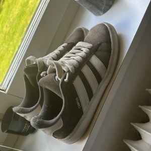 Adidas Campus sneakers grå/vita mocka - Säljer ett par klassiska Adidas Campus sneakers i grå mocka med vita detaljer och de ikoniska tre ränderna på sidorna. Skorna har vit sula, rund tå och snörning. Perfekta för dig som gillar retrostil och streetwear. Är inte använda så många gånger går att tvätta för en ny look.
