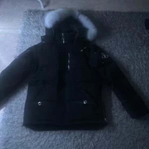 Svart dunjacka Moose Knuckles med pälshuva - Säljer en svart dunjacka från Moose Knuckles med ikonisk logga på ärmen och stor huva med ljus päls. Jackan har dragkedjor i sidorna, flera fickor med tryckknappar och snygga metalldetaljer. Perfekt för kalla vinterdagar och riktigt street. Priset kan diskuteras.