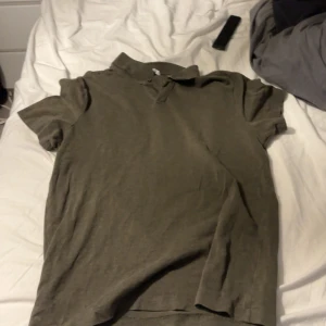 Olivgrön pikétröja med krage - M men passar S Olivgrön pikétröja med klassisk krage och korta ärmar. Tröjan har en enkel design utan synliga loggor eller mönster, perfekt för en clean och avslappnad look. Tillverkad i mjukt bomullstyg som känns skönt mot huden.