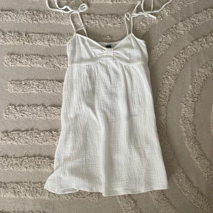 Vit sommarklänning från SHEIN - Superfin vit kortklänning från SHEIN i storlek XS. Klänningen har tunna axelband med knyt, lätt rynkad byst och smockad rygg. Tillverkad i mjuk bomull med diskret rutstruktur, perfekt för varma dagar.