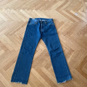 Levi's 501 blå jeans med råa kanter - Klassiska Levi's 501 jeans i blå denim med raka ben och råa, fransiga benslut. Jeansen har fem fickor, knappgylf och den ikoniska läderpatchen bak i midjan. Perfekt för dig som gillar en tidlös och avslappnad look.