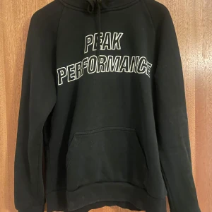 Svart hoodie från Peak Performance - Svart hoodie från Peak Performance med stor vit logga broderad över bröstet. Klassisk känguruficka framtill, snörning i huvan och långa ärmar. Perfekt för en chill och sportig look.
