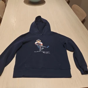 Mörkblå hoodie Polo Bear Ralph Lauren - Mörkblå hoodie från Polo Ralph Lauren med ikoniskt Polo Bear-tryck där björnen spelar hockey. Klassisk känguruficka framtill, huva och långa ärmar. Skön och mjuk, perfekt för kyliga dagar. Snygg streetstil med sportig vibe.
