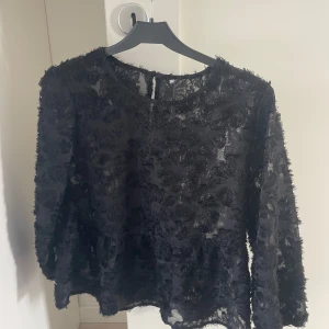 Svart transparent blus med blommig struktur - Säljer en svart transparent blus med trekvartslång ärm och blommig struktur i tyget. Blusen har rund halsringning och knäppning bak i nacken. Materialet är lätt och ger en luftig känsla, perfekt för att skapa en cool och unik look.