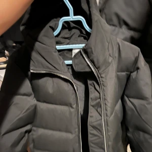 Svart lång pufferjacka från Grunt passar S-M  - Svart lång pufferjacka från Grunt med huva och markerade sömmar. Jackan har en rak passform, dragkedja framtill och är perfekt för kalla dagar. Enkel och stilren design som funkar till det mesta. Storlek XL/16Y. junior storlek.Köptes förra vintern men blev aldrig använd  nypris 1699 kr