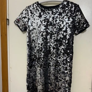 Glittrig paljettklänning från Bik Bok XS - Supersnygg kort klänning från Bik Bok i svart med massor av silvriga paljetter. Modellen är rak med korta ärmar och rund halsringning. Perfekt för dig som vill sticka ut med en glittrig look. Materialet är polyester och klänningen har en cool partyvibe.
