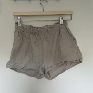Beige linneshorts från Lindex - Snygga beige shorts från Lindex i mjukt linnematerial. De har hög midja med resår och snörning samt två sidofickor. Perfekta för varma dagar och ger en chill vibe med sin loose fit och enkla design.