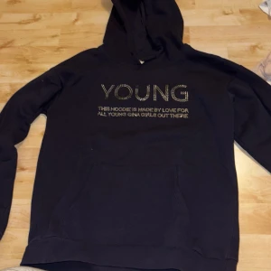 Svart hoodie från Gina Tricot med text - Svart hoodie från Gina Tricot med texten 'YOUNG' i glittriga stenar på bröstet. Klassisk känguruficka framtill och huva. Mjuk och skön bomullsblandning, perfekt för chill dagar. Snygg och enkel att matcha med jeans eller leggings.