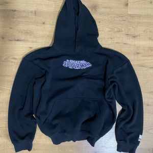 Svart Spiderman hoodie från H&M - Svart hoodie från H&M med relaxed fit och coolt Spiderman-tryck på bröstet samt en stor, färgglad Spiderman- och Venom-grafik på ryggen. Klassisk känguruficka och huva. Perfekt för dig som gillar Marvel och streetstyle.