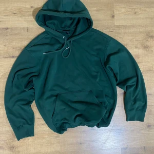 Mörkgrön hoodie från H&M loose fit - Mörkgrön hoodie från H&M i loose fit-modell. Tröjan har huva med snörning, magficka och ribbade muddar vid ärmslut och nederkant. Perfekt för en chill och avslappnad stil. Materialet är mjukt och skönt, troligtvis bomullsmix.