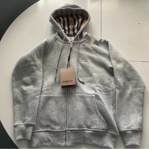 Burberry kofta - Säljer en grå hoodie från Burberry med klassiskt randigt foder i huvan. Tröjan har dragkedja framtill, snörning vid huvan och två stora fickor. Perfekt för dig som vill ha en stilren och bekväm look med premiumkänsla.