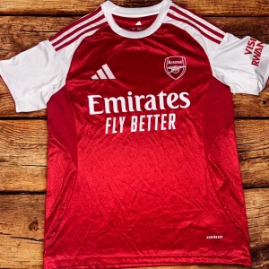 Arsenal hemmatröja  - Säljer en Högkvalitativ officiell Arsenal hemmatröja från Adidas i storlek S. Tröjan är röd med vita ärmar och detaljer, har klubbmärke och sponsortryck på bröstet samt klassiska Adidas-ränder på axlarna. Tillverkad i lätt och ventilerande material, perfekt för match eller träning.