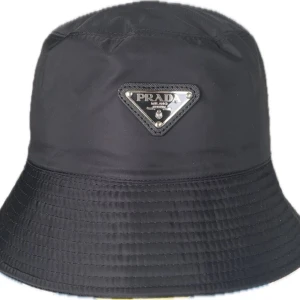 Svart bucket hat från Prada - Svart bucket hat från Prada med klassisk siluett och ikonisk metallogo framtill. Hatten är tillverkad i ett lätt och slitstarkt syntetmaterial som ger en clean och modern look. Perfekt accessoar för att lyfta din outfit med exklusiv streetstyle-vibe.