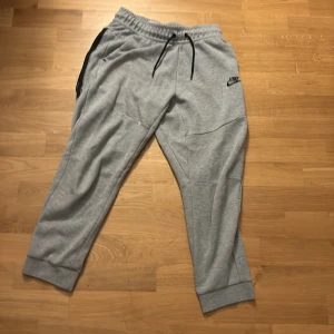 Grå mjukisbyxor från Nike  - Grå mjukisbyxor från Nike med svart logga på vänster ben. Byxorna har elastisk midja med svart snörning, ribbade muddar vid benslut och praktiska fickor med dragkedja. Perfekta för chill eller träning, tillverkade i mjukt bomullsmaterial.