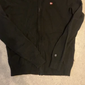 Svart kofta från Napapijri - Svart tröja från Napapijri med hel dragkedja framtill. Liten norsk flagga broderad på bröstet. Klassisk och stilren modell i stickat material, perfekt för lager-på-lager under kyliga dagar. Priset kan diskuteras och det är bara att ställa frågor vid behov. 