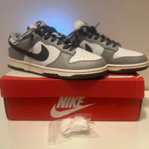 Nike Dunk Low light grey smoke - Nike Dunk Low sneakers i grått och vitt med svart swoosh och svarta skosnören. Klassisk låg modell med rund tå, perforerad tåbox och platt sula. Skorna är i syntet och skinn, levereras med extra vita skosnören och originalkartong.