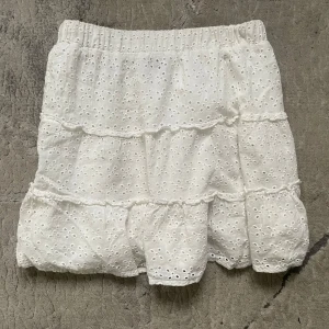 Vit volangkjol från FB Sister - Supersöt vit kortkjol från FB Sister med broderade blommor och volanger. Kjolen har resår i midjan och är gjord i ett lätt och luftigt bomullstyg, perfekt för varma dagar. Broderade detaljer ger en romantisk vibe och kjolen har en feminin siluett.