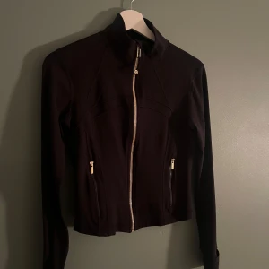 Svart cropped träningsjacka från Lululemon - Svart, figursydd cropped träningsjacka från Lululemon med guldig dragkedja framtill och på fickorna. Jackan har hög krage, långa ärmar och är tillverkad i ett stretchigt material av polyester och elastan. Perfekt för dig som gillar sportig och clean stil.