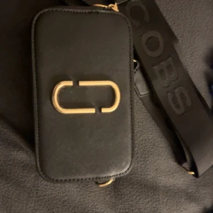 Svart axelväska från Marc Jacobs - Säljer en svart axelväska från Marc Jacobs med guldiga metalldetaljer och bred axelrem med logga. Väskan har två dragkedjor upptill och en ficka på baksidan. Stilren och praktisk modell i skinn med rektangulär form.