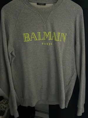 Grå sweatshirt från Balmain Paris - Säljer en grå sweatshirt från Balmain Paris med neongul logga framtill. Tröjan har rund halsringning och långa ärmar. Tillverkad i mjuk bomull för skön känsla och clean look. Perfekt för dig som gillar stilrena streetwear-plagg. Knappt använd