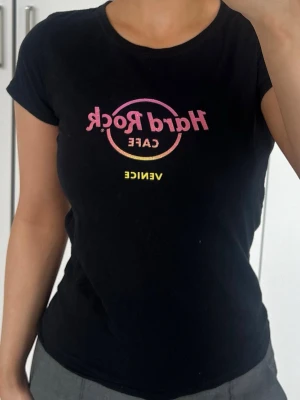 Hard Rock Cafe Venice svart t-shirt - Svart topp från Hard Rock Cafe Venice med tryck i rosa och gult framtill. Modellen är figurnära med rund hals och korta ärmar. Tillverkad i 100% bomull. Storlek L, tror det är barnstorlek, jag har XS/S och den passar mig 🖤