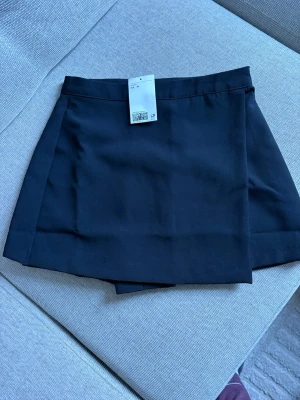 Svarta skorts från H&M, strl 34 - Svarta skorts från H&M i storlek 34. Klassisk omlott-look framtill med shorts under, vilket ger en snygg och bekväm stil. Gjorda i ett slätt tyg som känns mjukt och följsamt. Perfekt för dig som vill ha kjol-looken men ändå känna dig bekväm.