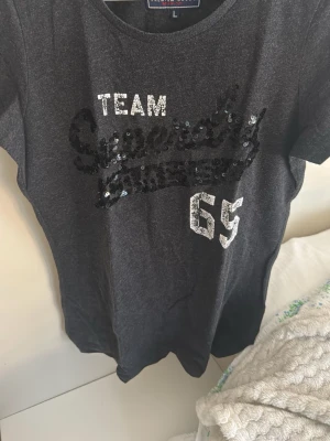 Mörkgrå Superdry t-shirt med paljetter - Snygg mörkgrå t-shirt från Superdry med texten 'TEAM' och siffran '65' i vitt samt svart paljettdekor framtill. Är storlek L men sitter mer som M/S. Klassisk rund hals och korta ärmar. Perfekt för dig som gillar sportig stil med lite extra bling.