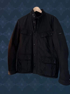 Barbour International Ariel Polarquilt jacka - Svart quiltad jacka från Barbour med klassisk ståkrage och mjukt foder. Jackan har flera fickor med lock och tryckknappar, samt dragkedja och knäppning framtill. Perfekt för kyliga dagar och snygg till de flesta outfits. •Nypris = Ca. 3499kr. • Priset går självfallet att diskutera vid en smidig affär. • Jackan är i mycket gott skick.