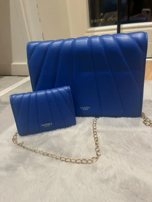 axelväska & plånbok - Säljer ett matchande set från Ulrika med en blå quiltad axelväska i syntetmaterial och tillhörande plånbok. Väskan har guldfärgad kedja och snygga diagonala sömmar. Plånboken har flera fack för kort och kontanter. Perfekt för dig som vill ha en färgklick i din outfit!