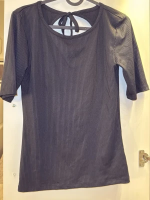 Svart ribbad topp med knyt i ryggen - Säljer en svart ribbad topp med halvlånga ärmar och snygg öppen rygg med knytband. Toppen har en rund halsringning och är gjord i ett stretchigt material som sitter bekvämt. 