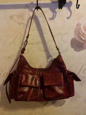 Vinröd axelväska  - Säljer en vinröd axelväska i skinn med justerbart axelband och flera stora fickor framtill och på sidorna. Väskan har dragkedja upptill och en cool, lite retro look. Perfekt för dig som vill ha plats för allt och ändå se stylish ut. Bara använd några få tal gånger så mycket fint skick inne och ute på väskan!