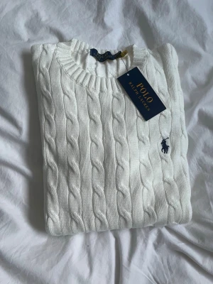 Vit kabelstickad tröja Polo Ralph Lauren - Vit kabelstickad tröja från Polo Ralph Lauren med klassisk rund halsringning och broderad marinblå logga på bröstet. Tröjan har ribbade muddar och är i mjuk bomull, perfekt för lager-på-lager under hösten och vintern.